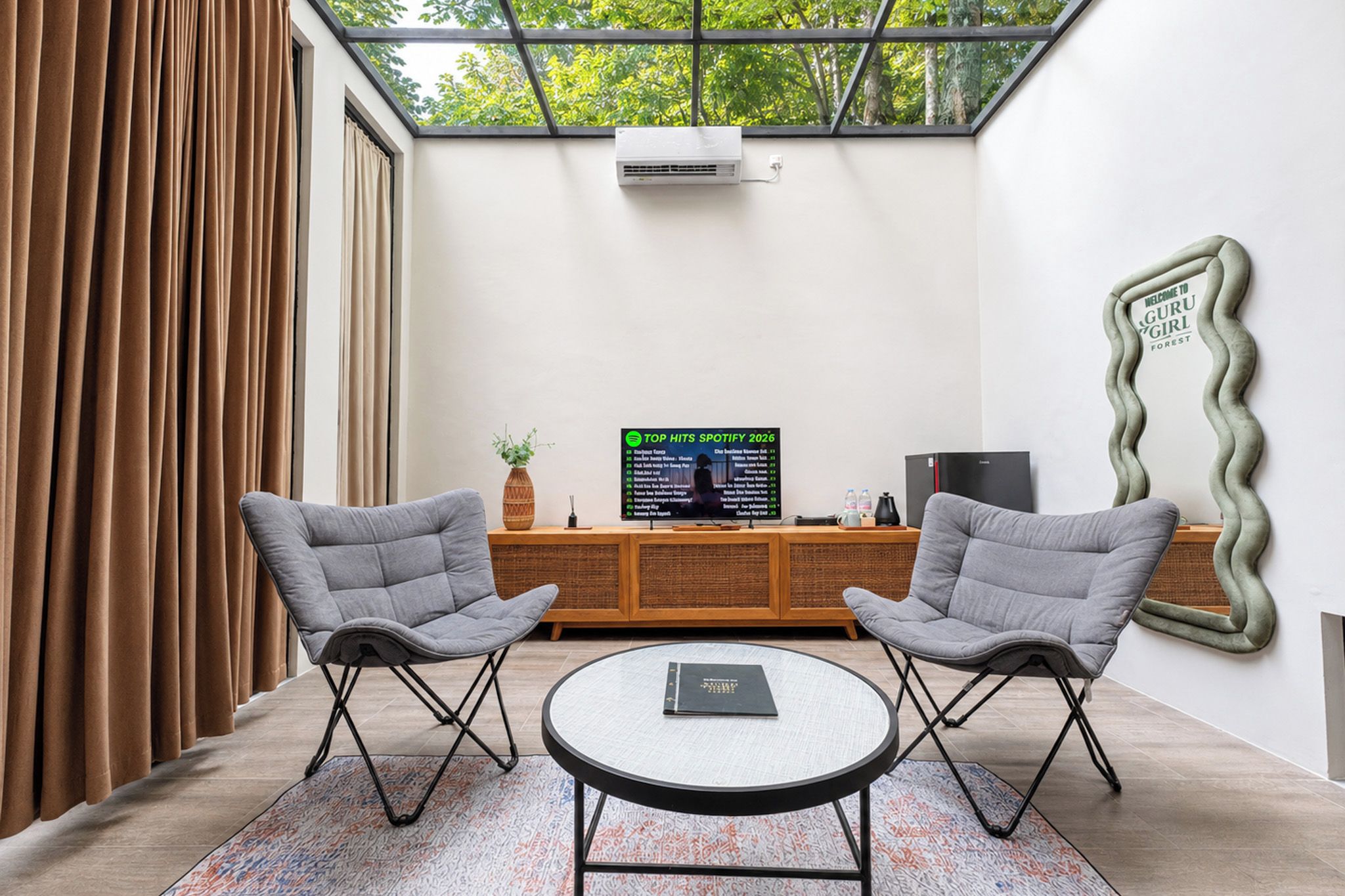 A modern room with a large window, two grey chairs, a small table, and a TV displaying a Spotify chart.