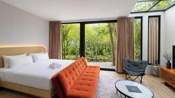 A modern hotel room with a large bed, a sofa, a chair, a small table, and a view of greenery through large windows.