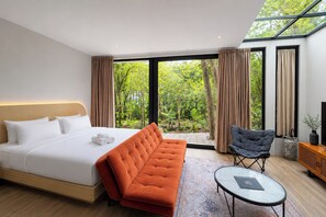 Ein modernes Hotelzimmer mit einem großen Bett, einer Couch, einem Sessel, einem kleinen Tisch und Blick auf Grün durch große Fenster.