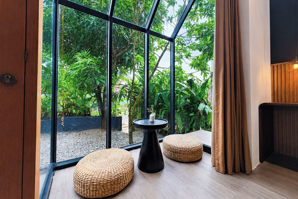 Un intérieur moderne avec de grandes fenêtres en verre, une table noire, deux poufs en fibres tressées et un plancher en bois.