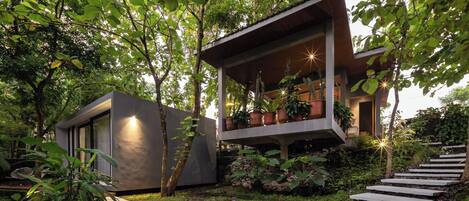 A modern house with a balcony, surrounded by lush greenery and a stone pathway.