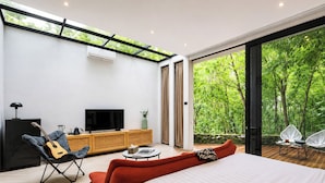 Quarto moderno com uma cama grande, televisão, um violão e vista para uma floresta exuberante.
