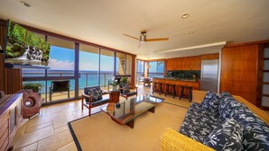 Een ruime woonkamer met een grote glazen wand met uitzicht op de oceaan, een keuken met houten kastjes en een eethoek met barstoelen.