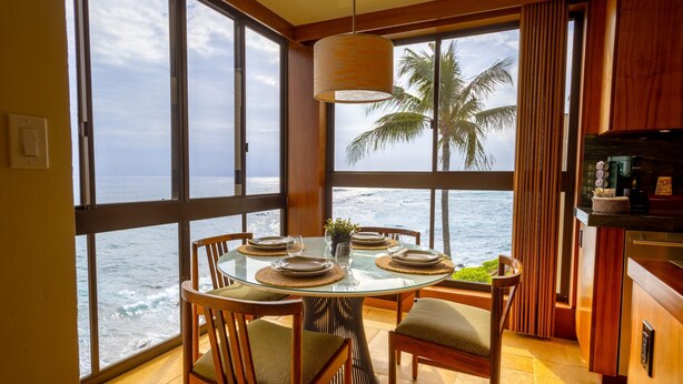 Een eetgedeelte met een glazen tafel en stoelen, uitzicht op de oceaan en een keuken met houten kasten.