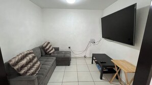 Een woonkamer met een grijze bank, een kleine tafel en een aan de muur bevestigde televisie.