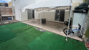 Een overdekt terras met een tafel, stoelen, een motor en een gazon van kunstgras.