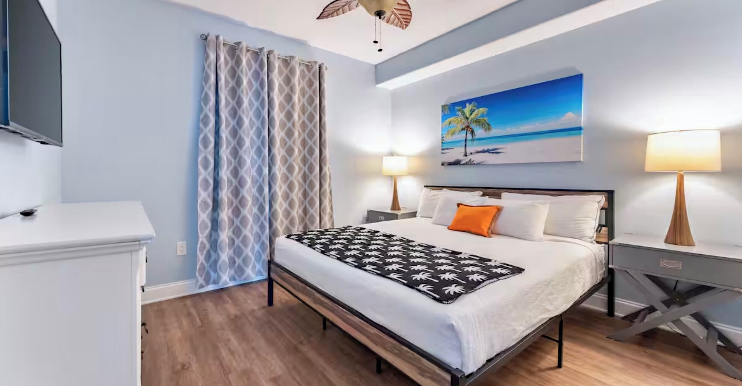 Een slaapkamer met een bed, nachtkastjes, een schilderij van een strand en een plafondventilator.