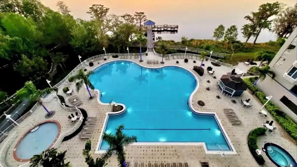 A large outdoor swimming pool with a unique shape, surrounded by lounge areas and a view of a lake at sunset. -  Sunrise lake view from the balcony. 3-bedroom condo near Disney/universal. (Orlando)