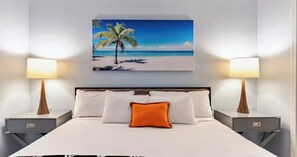 Ein Hotelzimmer mit einem Bett, zwei Nachttischen, einer Wandlampe und einem an der Wand hängenden Gemälde mit einer Strandansicht.