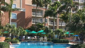 Piscina com escada de metal, cercada por palmeiras e um edifício com várias andares e sacadas.