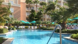 Piscina com espreguiçadeiras e guarda-sóis, cercada por palmeiras e um edifício de vários andares.