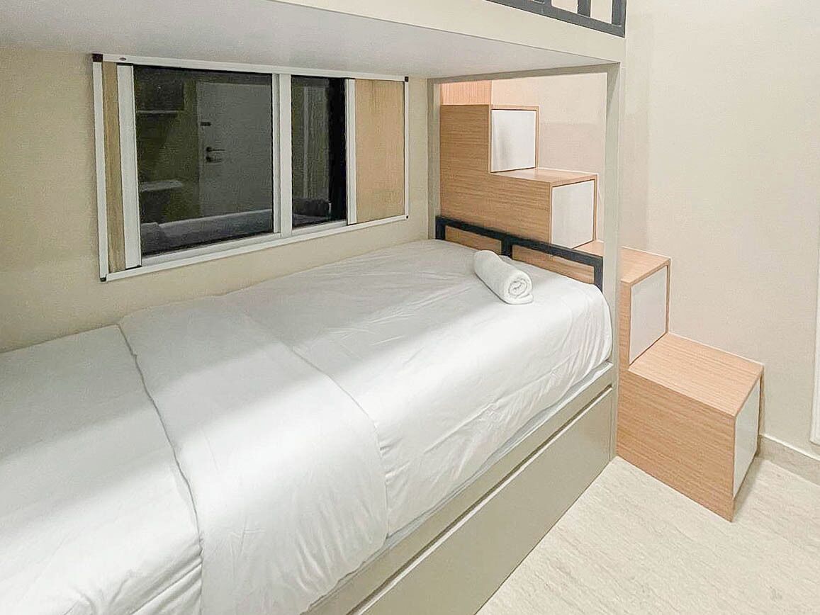 Un letto singolo con materasso bianco, persiane a una finestra e un piccolo comodino in legno.