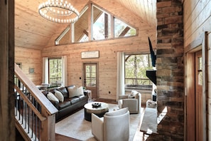 A wooden cabin interior with a staircase, a sofa, armchairs, a fireplace, and large windows. - New Build! 2B-2B Secluded Cabin/Hot Tub/Games/EV/Parking (Sevierville)