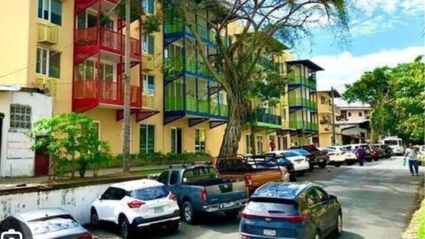 Un edificio residencial de varios pisos con balcones, coches aparcados y árboles.