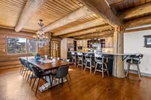 A spacious kitchen with wooden beams, a dining area with a large table and chairs, and a view of the outdoors. - Log Masterpiece with 5 Master Suites, Pool Table, Hot Tub (Silverthorne)
