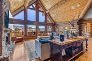 A log cabin interior with a fireplace, large windows, a wooden dining table, and a cozy seating area. - Log Masterpiece with 5 Master Suites, Pool Table, Hot Tub (Silverthorne)