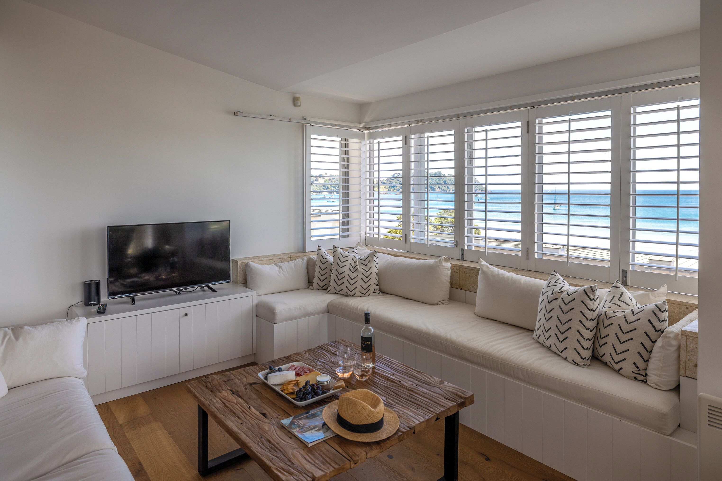 A modern living room with a large TV, a sofa with patterned cushions, a wooden coffee table with snacks and a bottle, and a view of the sea through large windows.