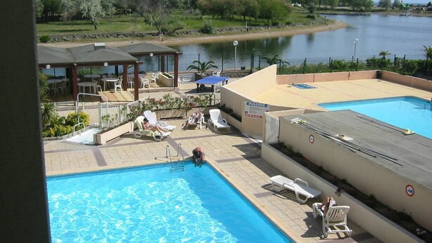 Piscina con sillones de descanso y un área cubierta, rodeada de un jardín ajardinado y una masa de agua.