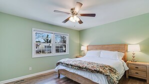 Ein Schlafzimmer mit einem hölzernen Bett, einem Deckenventilator, einem Nachttisch mit Lampe und einem Fenster mit Blick auf Bäume.