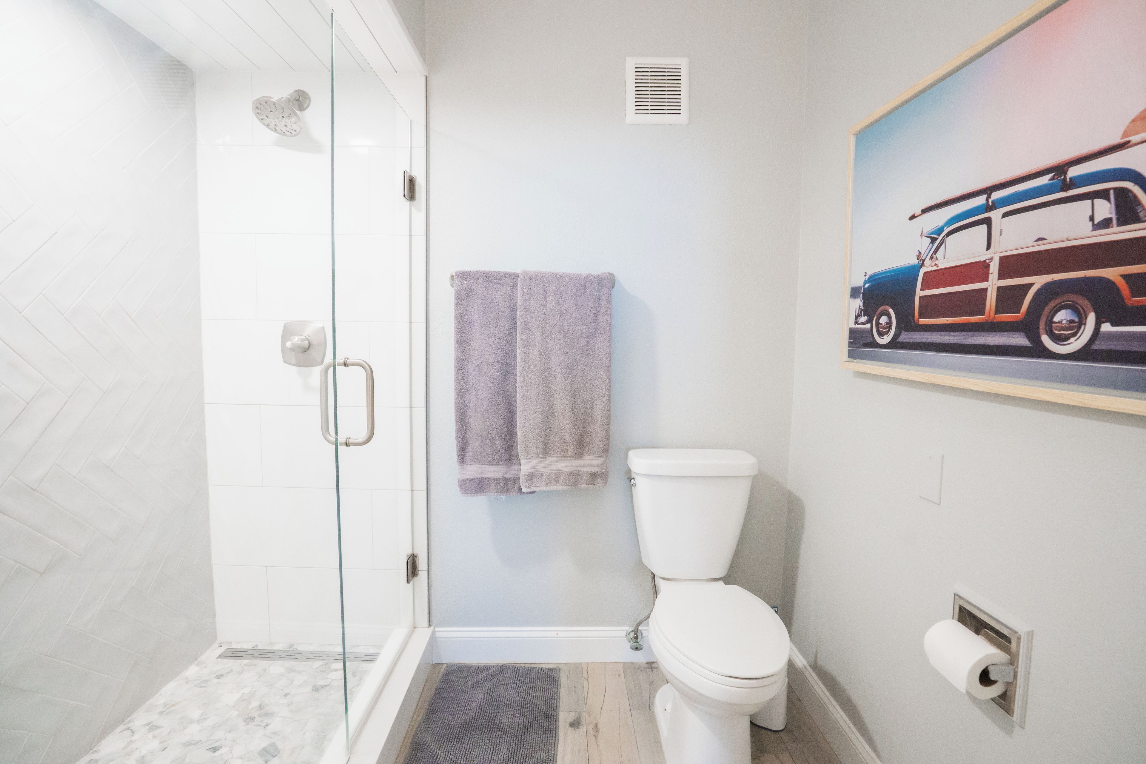 Un baño con cabina de ducha de vidrio, inodoro, toallero y un cuadro de un coche antiguo.