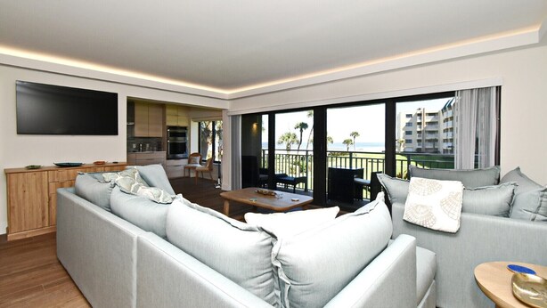 Ein modernes Wohnzimmer mit grauem Sofa, einem Flachbildfernseher, einer Küchenzeile und einem Balkon mit Blick auf Palmen und Gebäude.