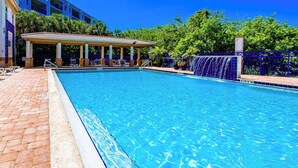 Una piscina con una zona de estar cubierta, una fuente de agua y un edificio al fondo.