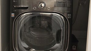 Uma máquina de lavar e secar dupla, compacta e em aço inoxidável, com painel de controle e uma garrafa de água vermelha no chão.
