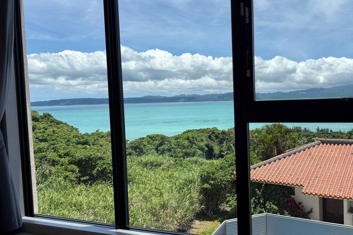 A view through a window of a coastal area with a building with a red-tiled roof and lush greenery.