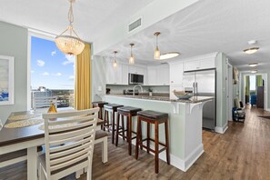 A modern kitchen with white cabinets, a stainless steel refrigerator, and a bar area with stools. - Fall Deals + Snowbird Rates | Gulf Front Beach Escape (Panama City Beach)