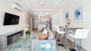 A modern living room with a flat-screen TV, a glass coffee table, a sofa, and a dining area with white chairs. - Romantic Island Retreat | Resort Pool & Sunset Rooftop (Battaleys)