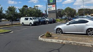 A parking lot with several cars, a large signboard with multiple business names, and a clear blue sky with some clouds.