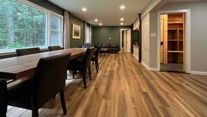 Una habitación amplia con una mesa de madera larga y sillas, un sofá, y vista a un bosque a través de ventanas grandes.