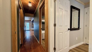 Un pasillo con pisos de madera y puertas blancas, que conduce a una habitación con un espejo grande y una puerta.