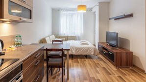 Habitación compacta y moderna con kitchenette, comedor, cama, televisor y ventana con cortinas translúcidas.