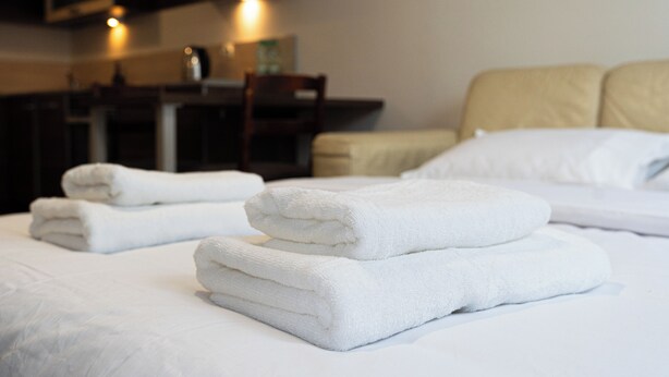 Habitación de hotel con toallas blancas impecables sobre la cama, una silla de madera y un escritorio con lámpara.