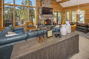 A log cabin interior with a stone fireplace, leather sofa, and large windows offering mountain views. - Skyfall Cabin | Luxury, Ski In/Out | Big Sky Resort (Big Sky)