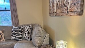 Un salón con un sofá gris, cojines estampados, una manta, una mesita de madera con una lámpara y una obra de arte colgada en la pared.