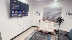 A modern living room with a flat-screen TV mounted on the wall, a glass coffee table, a white leather sofa, and a black leather couch. - Cozy 1-bedroom apartment in perfect Mandeville with AC and gym access (Mandeville)
