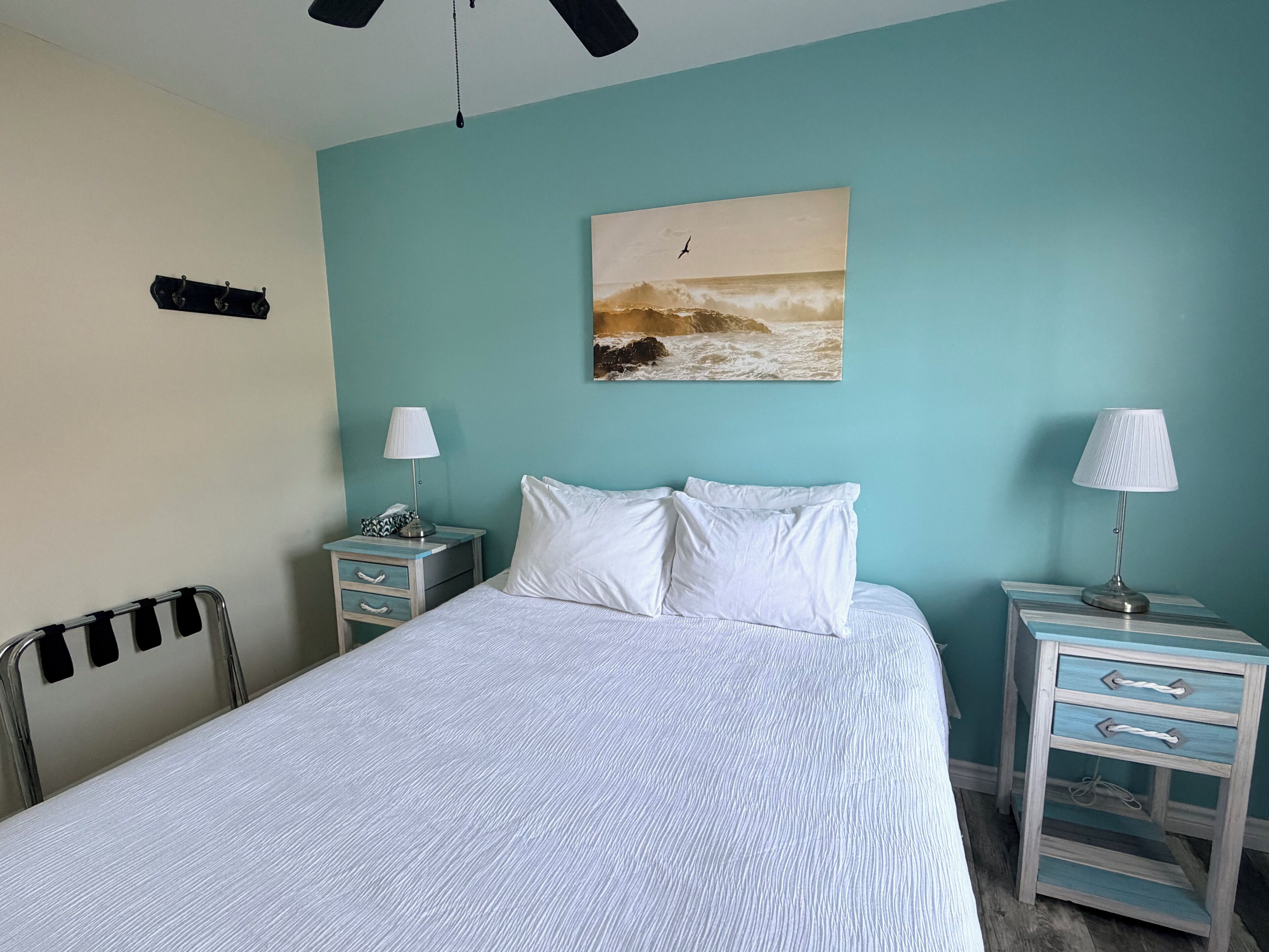Un dormitorio con una cama, dos mesitas de noche, una lámpara colgante, un cuadro marino y un ventilador de techo.