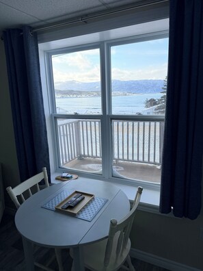 A room with a round table and chairs, a window with a view of a snowy landscape and a body of water. - Drift Ashore House (Rocky Harbour)