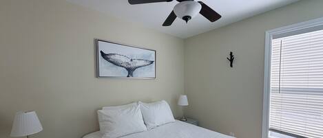 Un dormitorio con cama, ventilador de techo, pintura de cola de ballena en la pared, mesita de noche con lámpara y ventana con persianas.