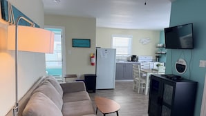 Een moderne woonkamer met een bank en een kleine tafel, een keuken met een koelkast en fornuis, en een televisie die aan de muur is bevestigd.