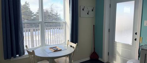 Ein Zimmer mit Esstisch, Stühlen, einem Fenster mit Vorhängen, einer Tür, einem Besen und einem Mülleimer.