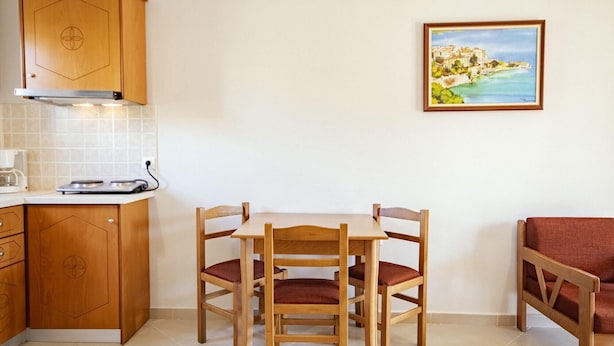 A compact kitchen with wooden cabinets, a dining table with four chairs, and a painting of a coastal scene on the wall. - Robolla Beach (Corfu)