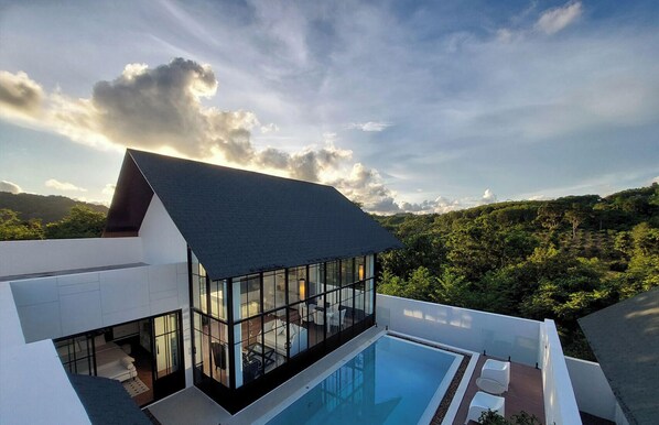 Una casa moderna con un amplio balcón de vidrio que da a una piscina y exuberante follaje.