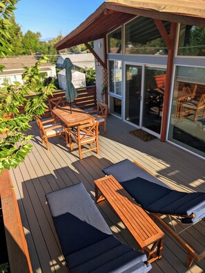 Una terraza de madera con una mesa y sillas, un espacio cubierto y un ventanal de vidrio que da a un ambiente interior.