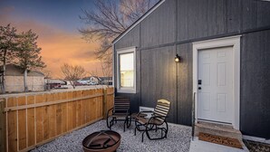 A house with a dark exterior, a white door, and a wooden fence. There are two chairs and a fire pit on the gravel patio. - Modern & Cozy Home Near Fort Sill (Lawton)