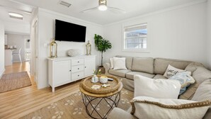 A modern living room with a beige sofa, a round coffee table with decorative items, a flat-screen TV mounted on the wall, and a ceiling fan. - Newly renovated cottage! Walk to the beach! (Fernandina Beach)