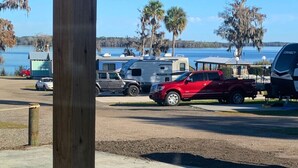 Ein Campingplatz mit Wohnmobilen und Autos, die am Seeufer geparkt sind, umgeben von Palmen und einem klaren Himmel.