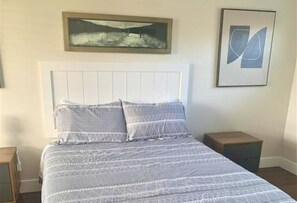 Ein Bett mit Kopfteil, ein Nachttisch, ein gerahmtes Bild über dem Bett und ein kleiner Tisch daneben.